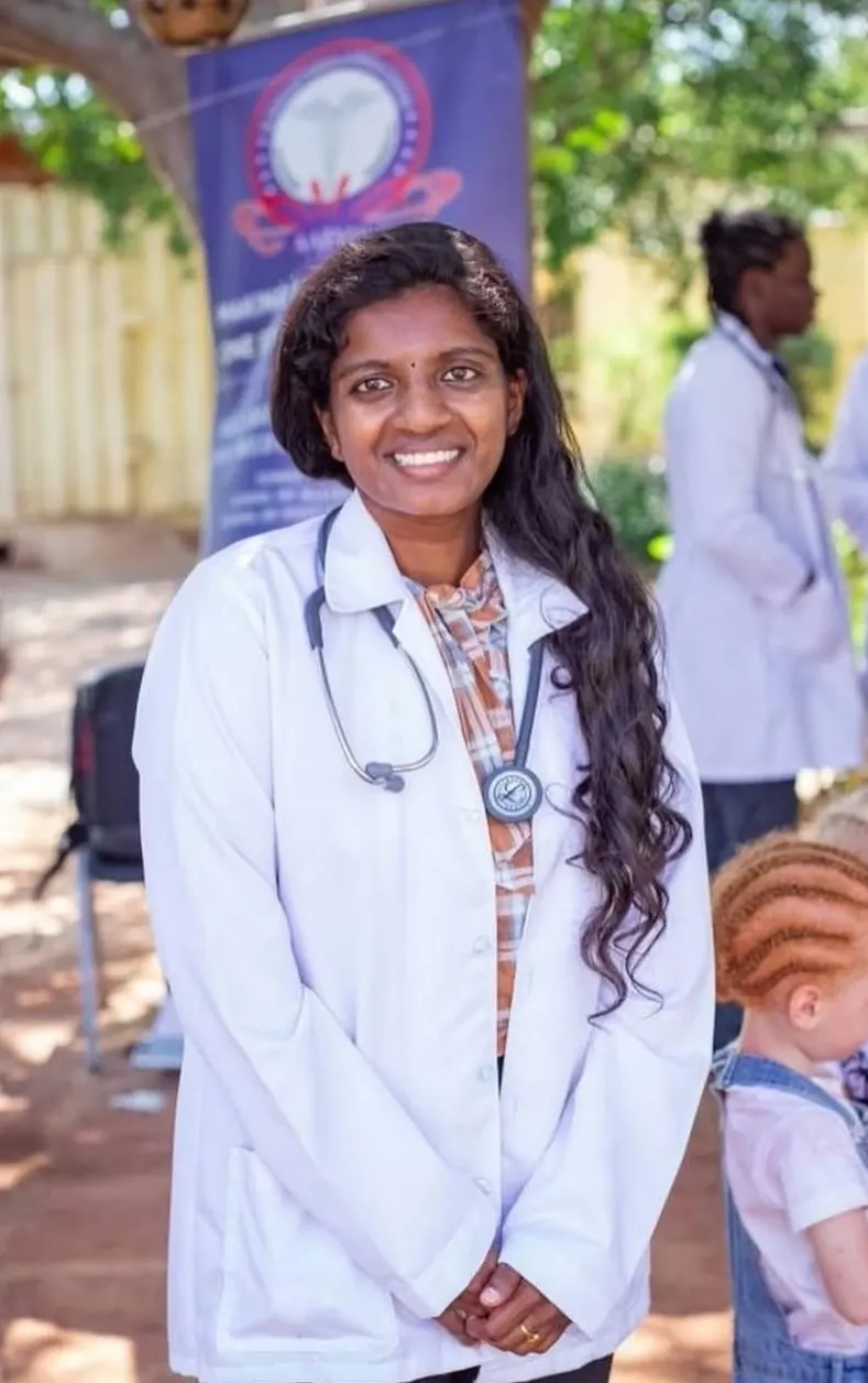An AAIMS student in a white coat with a stethoscope around her neck, smiling at the camera at an outdoor community health event.